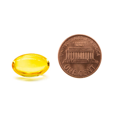 A clear yellow softgel capsule being compared to the size of a penny